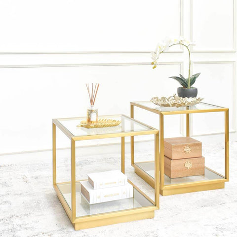 A matching pair of gold side tables with glass tabletops, one adorned with a golden leaf-shaped tray containing Finn Avenue reed diffuser. Both are placed on a luxury rug against a white living room feature wall.