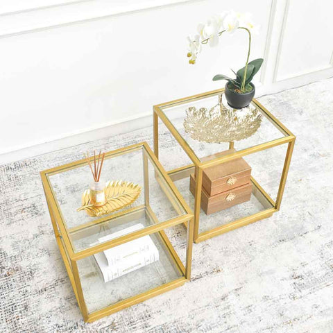 Two gold side tables with glass tabletops, one featuring a golden leaf-shaped tray and orchid plant, placed on a neutral coloured rug against a white wall.