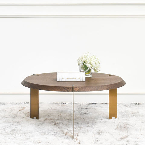 A modern coffee table featuring a circular top made from elm wood natural veneer, paired with three angular stainless steel legs plated in a bronze-gold finish. The table is styled with a white book and a small glass vase of white flowers, placed in a bright, minimalistic room with a neutral textured rug.