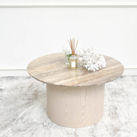 A round coffee table with a travertine marble top and a neutral-toned wooden base, styled with minimal decor, including a white coral centerpiece, a reed diffuser, and a glass vase with white flowers. The table is set on a light, textured area rug in a modern interior.