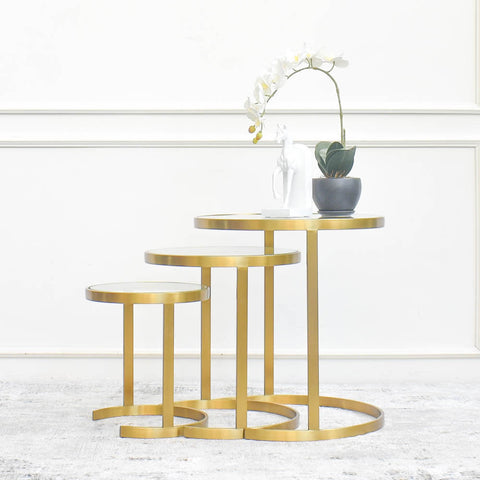 Three gold steel side tables with circular tiers, arranged in ascending order of height, with a white orchid plant and a decorative figurine on the tallest table. They are placed against a white wall with decorative paneling.